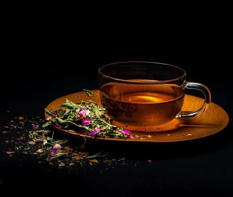 Tazza trasparente su un piattino in legno con un infuso di colore ambrato. Intorno erbe e piccoli fiori rosa scura . Il tutto su uno sfondo scuro. Ecco un ottimo modo per servire le tisane