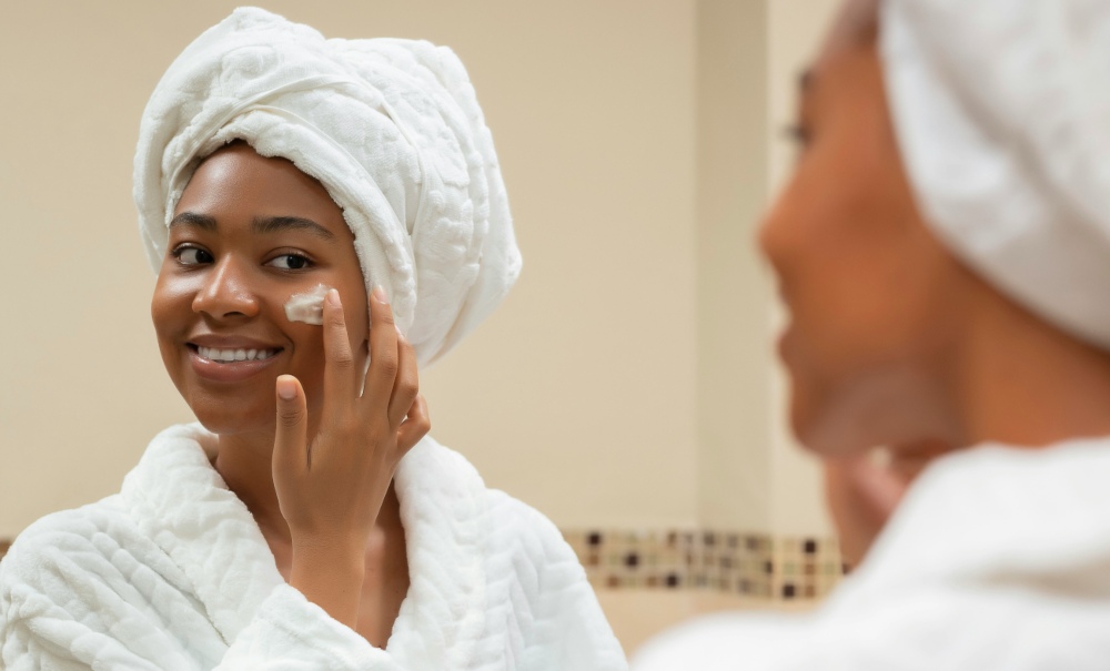 Donna in accappatoio bianco davanti allo specchio che spalma la crema sul viso per preparare la pelle del viso all'estate