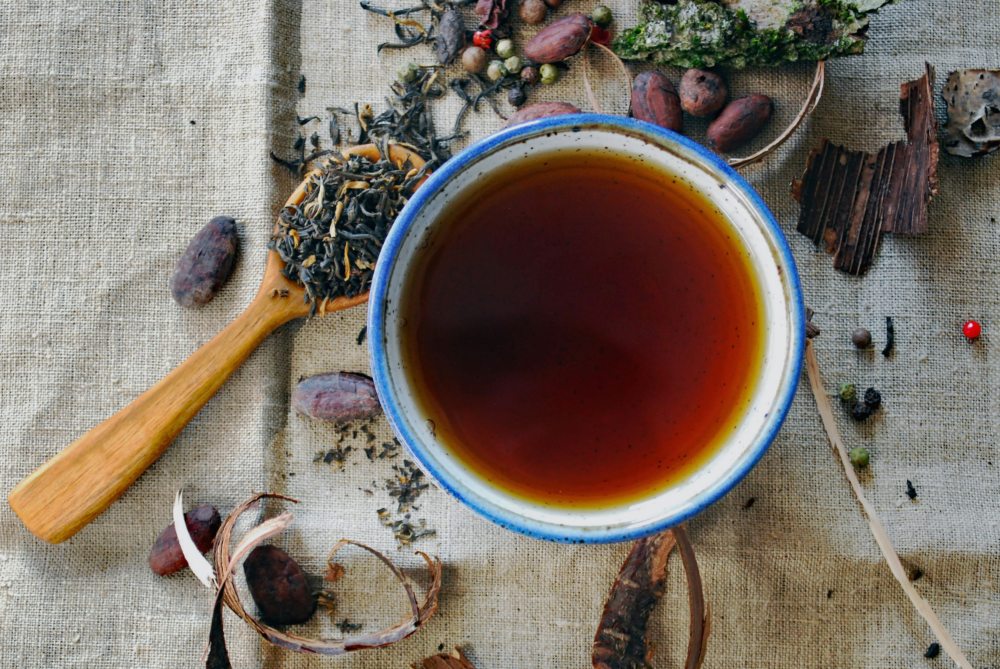 Prepararsi all'estate e alla prova costume: una tazza di tisana viasta dall'alto su tovaglia di lino grezzo con fiori, bacche e foglie essiccate spese e un cucchiaino in legno