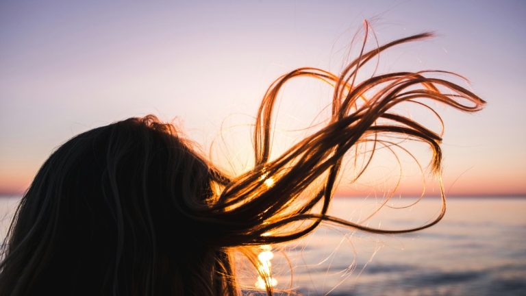 Dettaglio di donna di spalle con lunghi capelli scuri liberi dalla secchezza del cuoio capelluto. Sullo sfondo un tramonto estivo sul mare